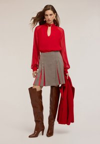 Blusa rossa trasparente a maniche lunghe con scollatura a occhiello, gonna plissettata tartan, stivali marroni al ginocchio e un cappotto rosso appoggiato sul braccio.