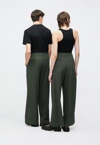 Παντελόνια με φαρδύ πόδι σε σκούρο πράσινο ύφασμα, με ψηλή μέση, πίσω τσέπες και λεία υφή, συνδυασμένα με μαύρα τοπ και χοντροκομμένα παπούτσια.