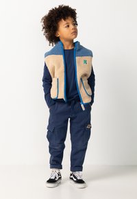 Kind mit lockigem Haar steht mit den Händen in den Taschen, trägt eine tanfarbene und blaue Fleeceweste, ein marineblaues Langarmshirt, marineblaue Cargo-Hosen und schwarze Sneaker.