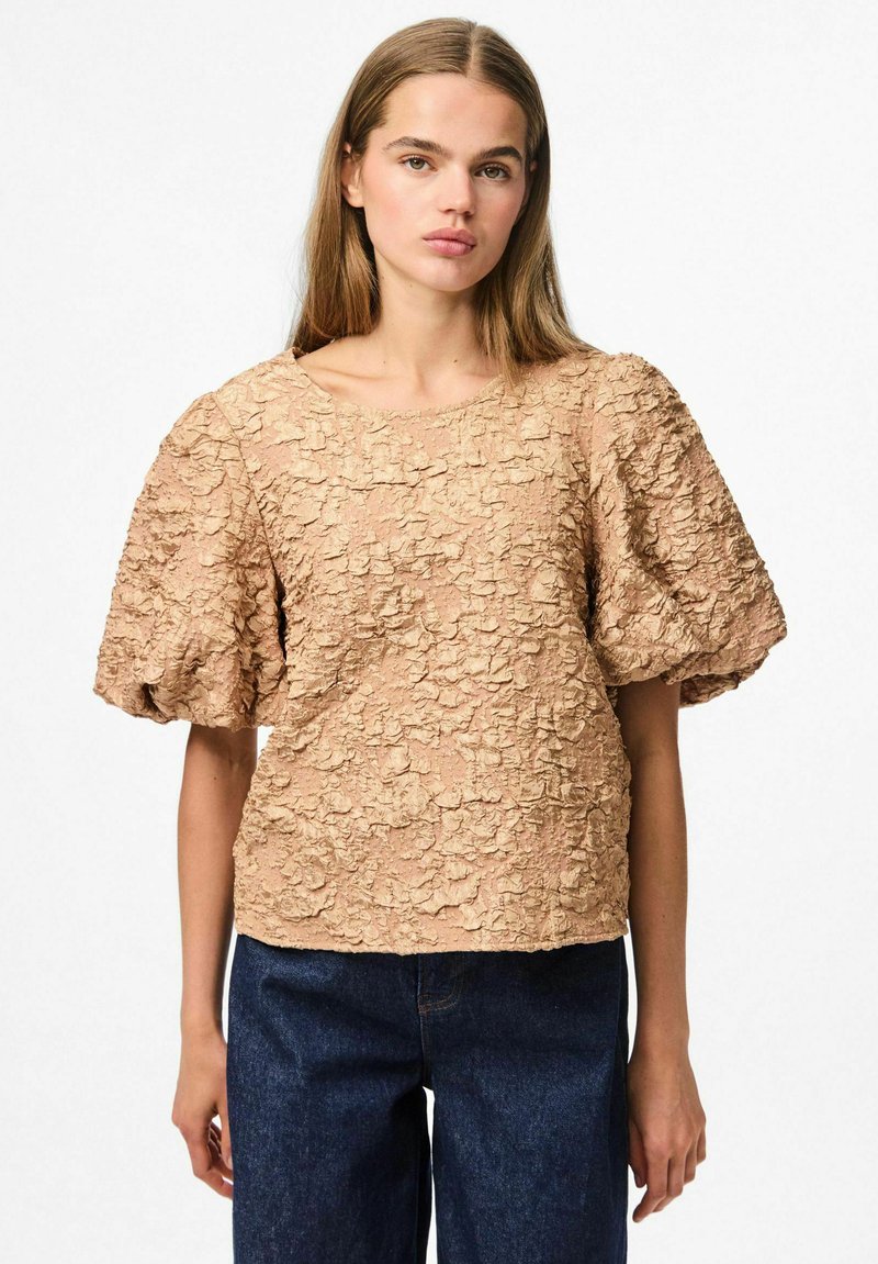 Top beige con texture e maniche corte a sbuffo, con un motivo floreale e un collo rotondo. Abbinato a jeans di denim blu scuro.