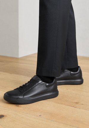 Osoba nosząca czarne skórzane sneakersy sznurowane i czarne spodnie stoi na jasnej drewnianej podłodze przy białej ścianie.