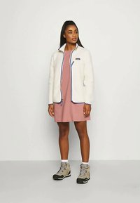 Bílá fleecová bunda s modrými detaily, předním zipem a logem. V kombinaci se světle růžovými šaty a béžovými turistickými botami s černými podrážkami.