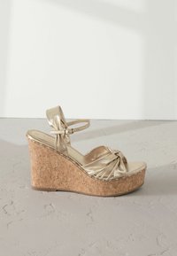 Zlaté metalické wedges sandále s korkovou platformou, s uzlovým dizajnom na hornej časti a nastaviteľným členkovým remienkom pre lepšie prispôsobenie.