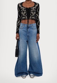 Femme portant un haut court noir à manches longues avec des lignes abstraites blanches, un jean bleu à jambes larges, des chaussures argentées, et tenant un petit sac à main noir.