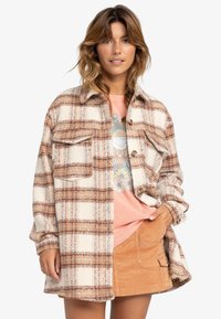 Geruite shirtjack in crème, bruin en lichtblauw. Zacht, textuurstof met twee borstzakken en knoopsluiting aan de voorkant. Draagt over een roze shirt.
