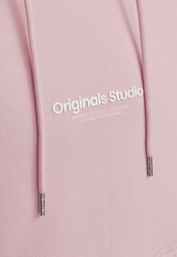Gai�ši rozā kapucis ar regulējamiem aukliņām, kuram ir baltais logo "Originals Studio" un sudraba aksesuāri uz aukliņām.