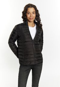 Sort puffer-jakke med lynlås, quiltet tekstur, høj krave og sidelommer, båret over en hvid sweater og parret med mørkeblå jeans.