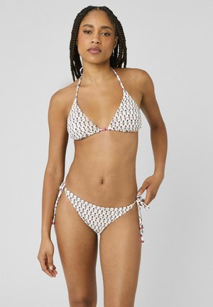 Jeune femme aux cheveux tressés portant un bikini à imprimé floral blanc à col halter, ajustant le lien du bas du bikini devant un fond uni.