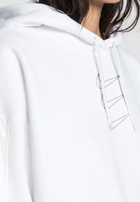 Vit huvtröja i sweatshirtmaterial med stor luva. Har en minimalistisk svart grafik och dragsnören för justering.