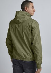 Giacca con cappuccio verde oliva, con polsi e orlo elasticizzati. Realizzata in tessuto leggero e liscio; presenta una vestibilità comoda.