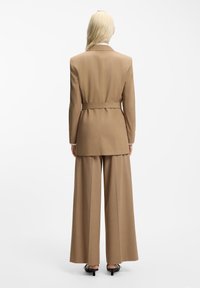 Μπεζ κοστούμι με ζώνη στη μέση. Διαθέτει παντελόνια με φαρδιά πόδια και μακρύ σακάκι με άνοιγμα στο πίσω μέρος. Λεία υφή ύφασμα με δομημένη σιλουέτα.