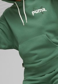 Zielona bluza z kapturem wykonana z miękkiego materiału, posiadająca kieszeń kangurzą, białe sznurki do ściągania oraz haftowane logo Puma na klatce piersiowej.