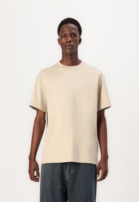 Beige Baumwoll-T-Shirt mit rundem Halsausschnitt, kurzen Ärmeln und einer entspannten Passform. Tragen Sie es mit dunkelblauen Denim-Hosen, die ein einfaches Design zeigen.