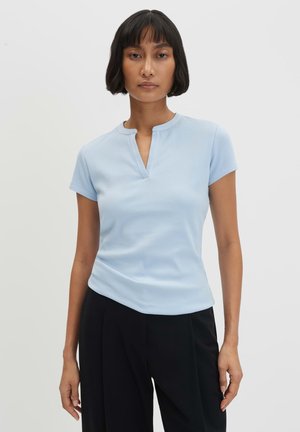 Frau trägt ein hellblaues, geripptes Kurzarmoberteil mit kleinem V-Ausschnitt und schwarze hochtaillierte Hose vor einem schlichten Hintergrund.