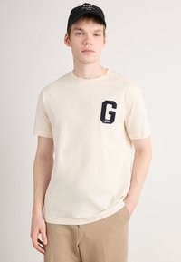 Jeune homme portant un t-shirt beige avec un emblème noir en forme de "G" sur la poitrine et une casquette noire, se tenant devant un fond clair uni.