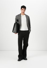 Hall, hallid blazer valge särke peal, mustad laiad püksid, hoides musta tekstureeritud käekotti ja jalas mustad kingad. Minimalistlik stiil valge taustal.