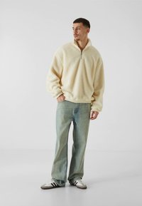 Pulover iz kremastobele flis tkanine s četrtinskim zadrgo in teksturirano površino. Kombiniran s svetlo modrimi kavbojkami in rjavo obutvijo.