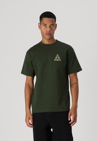 T-shirt en coton vert foncé avec un col rond et des manches courtes, présentant un logo triangulaire jaune sur la poitrine gauche.