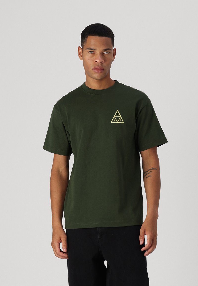 Camiseta de algodón verde oscuro con cuello redondo y mangas cortas, que presenta un logo triangular amarillo en el lado izquierdo del pecho.