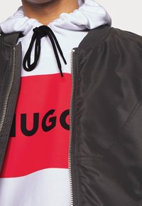 Dunkelgraue Bomberjacke mit einem silbernen Reißverschluss, kombiniert mit einem weißen Hoodie, der ein auffälliges rotes Grafikdesign und schwarzen Text "HUGO." zeigt.