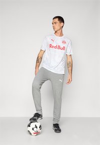 White short-sleeve shirt with marble texture and red "Red Bull" logo, paired with gray sweatpants and black athletic shoes on a soccer ball.