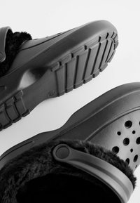 Schwarze Clogs mit strukturierter Oberfläche, die über Belüftungsöffnungen und ein weiches Innenfutter verfügen. Robuste Gummisohle mit ausgeprägtem Profil.