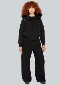 Chaqueta acolchada negra con capucha forrada de piel, cierres de cremallera y botones, grandes bolsillos delanteros, combinada con pantalones anchos negros y zapatillas.