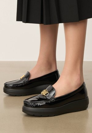 Fényes fekete platform talpú mokaszin arany emblémával, fekete pliszírozott szoknyával viselve, semleges alapon, padlón bemutatva.