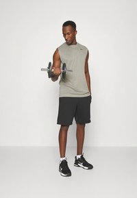 Grijs mouwloos sportshirt, zwarte short, een metalen dumbbell vasthoudend. Zwarte sportschoenen met witte accenten. Eenvoudige achtergrond.