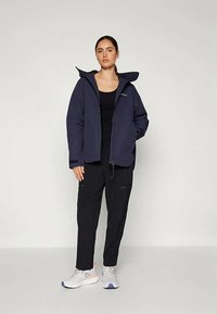 Dark navy waterproof jacket with a front zipper, adjustable hood, and two side pockets, paired with black pants and sneakers.
