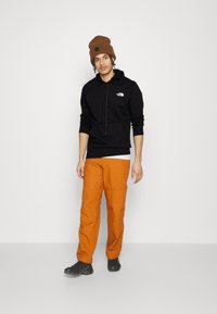 Felpa nera con cappuccio e logo, pantaloni cargo arancioni e calzature nere. Berretto lavorato a maglia marrone. Look semplice e casual da streetwear.