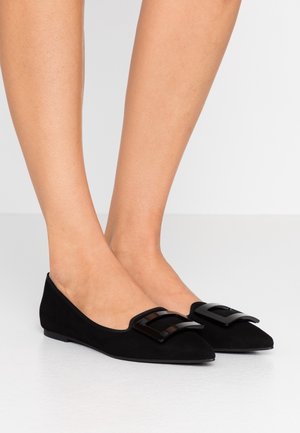 Jambes féminines portant des ballerines noires à bout pointu avec un grand détail de boucle noire, debout sur un sol blanc réfléchissant.