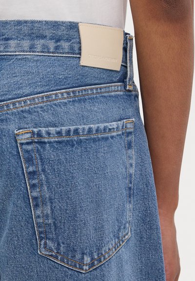 Helle blaue Denim-Jeans mit glatter Textur, versehen mit einer Gesäßtasche und einem beigen Lederpatch mit geprägt eingestanztem Branding am Bund.