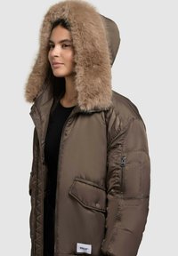 Puffer jacket in dark brown with a large faux fur-lined hood, zip pockets on sleeves, and a white logo patch at the waist.
