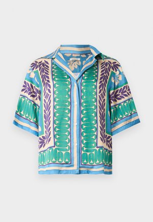 Vrouw met lang zwart haar, gekleed in een kort mouwig patroonhemd en brede beige broek, die een kleine zwarte handtas met een gouden ring vasthoudt.