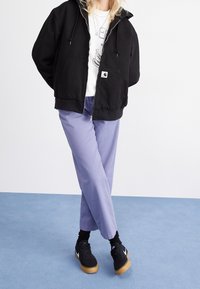 Black zip-up hoodie, white graphic t-shirt, light purple pants, black sneakers with gum soles, set against a plain background.