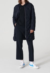 Tikkitettu tummansininen pitkä takki, jossa on huppu, yhdistetty mustaan villapaitaan ja tummaan housuun. Valkoiset tennarit sinisillä yksityiskohdilla täydent�ävät asukokonaisuutta.