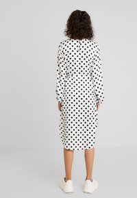 Langärmliges weißes Kleid mit großen schwarzen Punkten, tailliertem Schnitt und einem Schlüssel-ausschnitt im Rücken. Getragen mit hellen Sneakers.