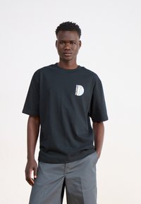 Black cotton t-shirt with short sleeves, featuring a small white graphic logo on the chest. Paired with light gray, relaxed-fit pants.