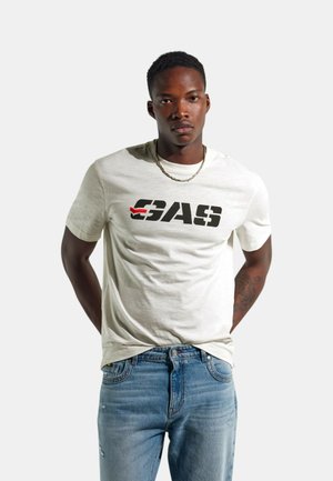 Giovane uomo che indossa una t-shirt bianca con il logo GAS, jeans azzurri chiari, collana d'oro, in piedi con le mani dietro la schiena su uno sfondo semplice.