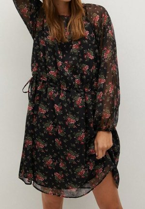 Femme portant une robe longue noire transparente à manches longues avec des motifs floraux roses et verts, cintrée à la taille par un lien latéral.