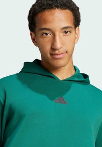 Zelená mikina s kapucí z měkké látky, vybavená přední kapsou, kapucí se šňůrkami a jemným černým logem Adidas na hrudi.