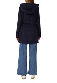 Marineblauer Wollmischungs-Mantel mit Kapuze, tailliertem Gürtel und knöpfbaren Manschetten, kombiniert mit weit geschnittenen Jeans und weißen Sneakern.
