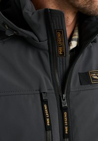 Black softshell jacket with zippered pockets, orange and black branding labels, and a high collar. Smooth texture, practical design.