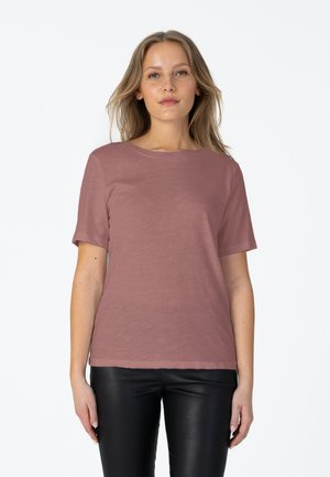 Jeune femme aux longs cheveux blonds porte une chemise mauve à manches courtes et un pantalon en cuir noir, debout sur un fond blanc uni.