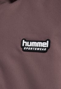 Schwarz-weißer Hummel-Sportbekleidungs-Patch auf einem strukturierten mauvefarbenen Stoffhintergrund.