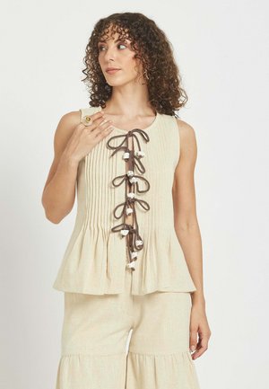 Ženska s kodrastimi lasmi, oblečena v brezrokavnik bež barve z rjavimi pentljami in krem hlače z nabranim robom, stoji pred enobarvnim ozadjem.