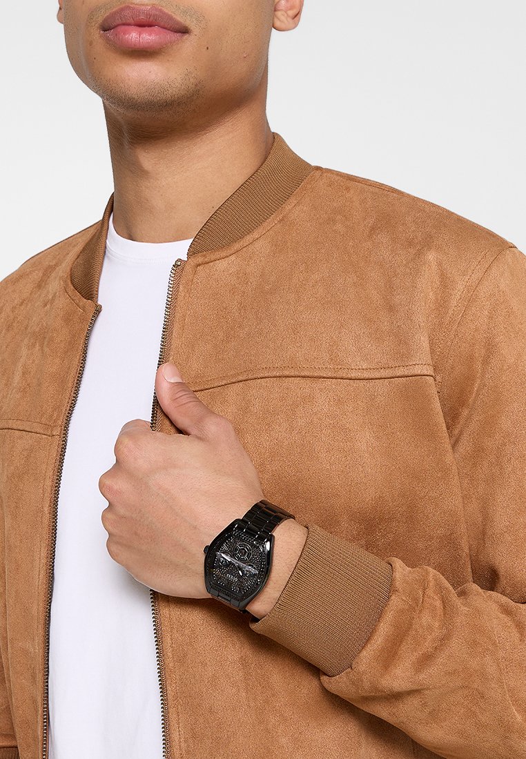 Hombre con una chaqueta de ante color beige sobre una camisa blanca, mostrando un reloj de pulsera de metal negro en su muñeca izquierda.