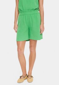 Zelena gubasta krpna kratka hlače z elastičnim pasom, ki imajo sproščen kroj in stranske žepke. Obuvene s svetlima tkaninami brez vezalk.