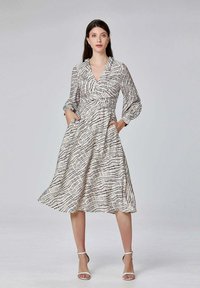 Robe à manches longues blanche avec un motif en croix noir, décolleté en V et poches latérales. Tissu fluide et longueur mi-mollet.